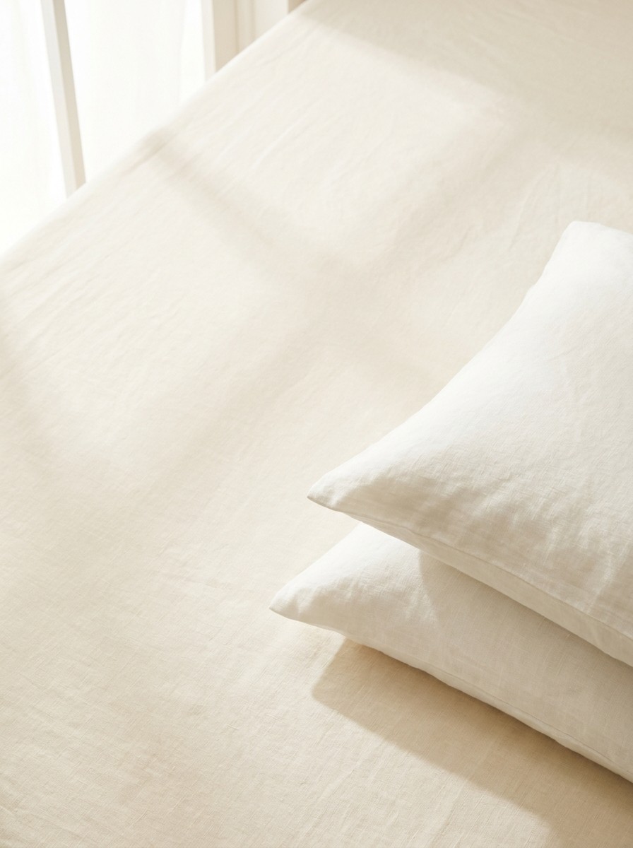 Two stacked white linen pillows pushed to the lower-right of cream bedding, the upper-left left empty in soft window light.