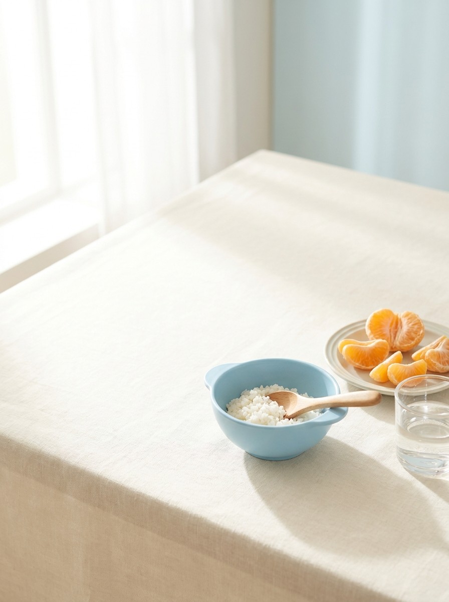 A sky-blue children's rice bowl with a wooden spoon, a small plate of peeled tangerine segments and a glass of water on a cream linen tablecloth, with open space at upper left.