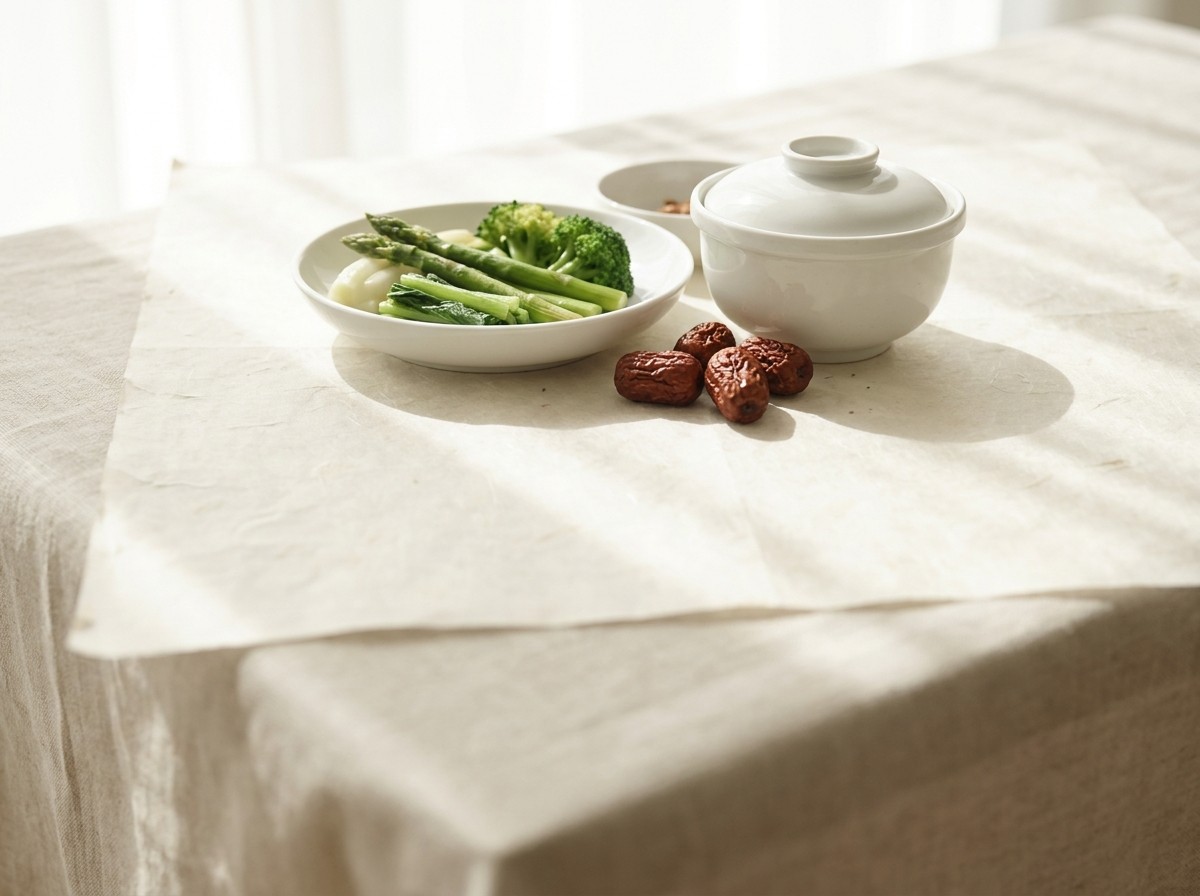 A white porcelain dish of blanched greens, a covered rice bowl and a few dried jujubes grouped on cream hanji paper in soft window light, with open surface below.