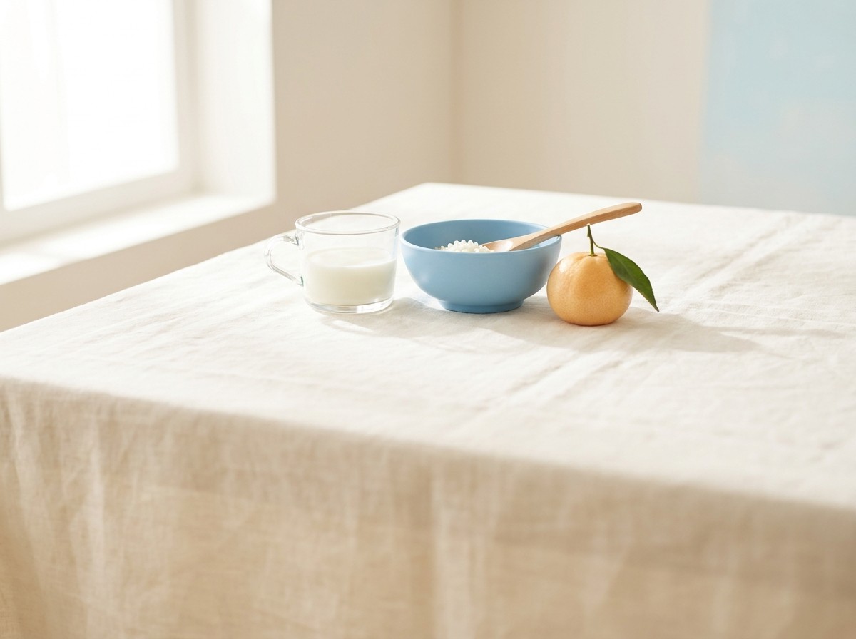 A small clear glass cup of milk, a sky-blue children's rice bowl with a wooden spoon and a whole tangerine with a leaf grouped on a cream linen tablecloth in soft window light.