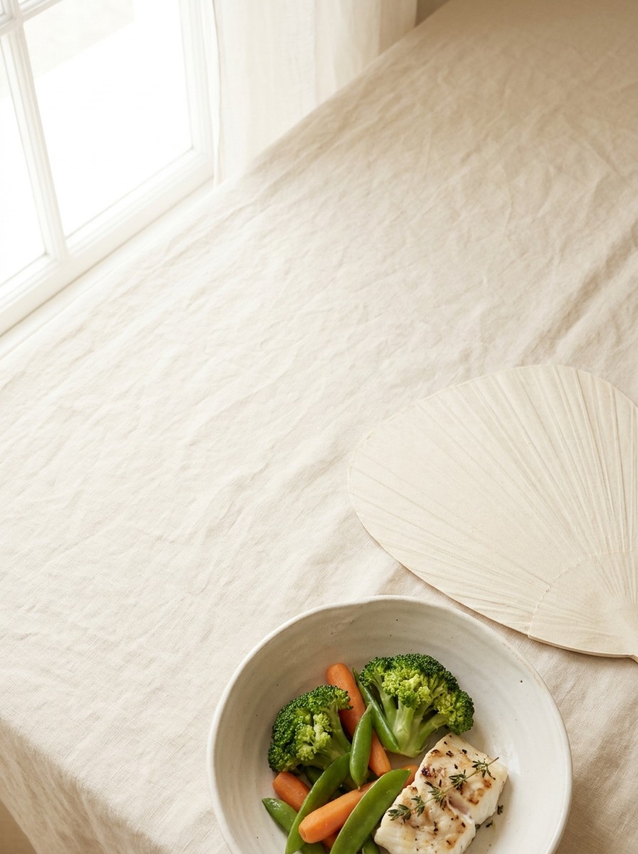 A pale paper hand fan and a ceramic bowl of steamed vegetables with grilled fish pushed to the lower-right of cream linen, the upper-left left empty in window light.