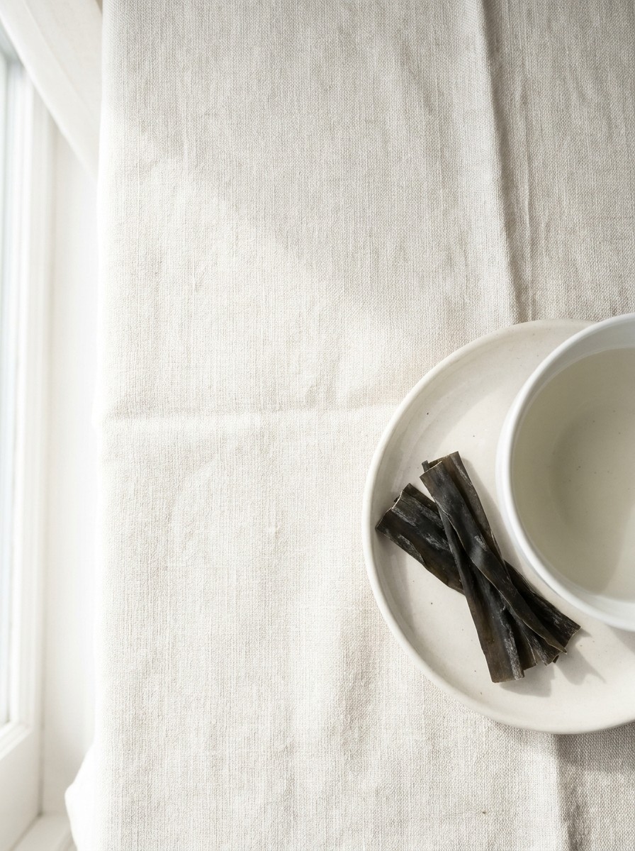 A small white porcelain dish of dried kelp strips beside a white bowl pushed to the lower-right of cream linen, the upper-left left empty in soft window light.