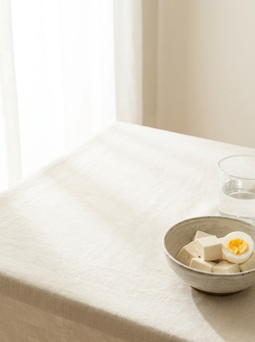 A ceramic bowl of tofu cubes with a halved boiled egg and a glass of water pushed to the lower-right of cream linen, the upper-left left empty.