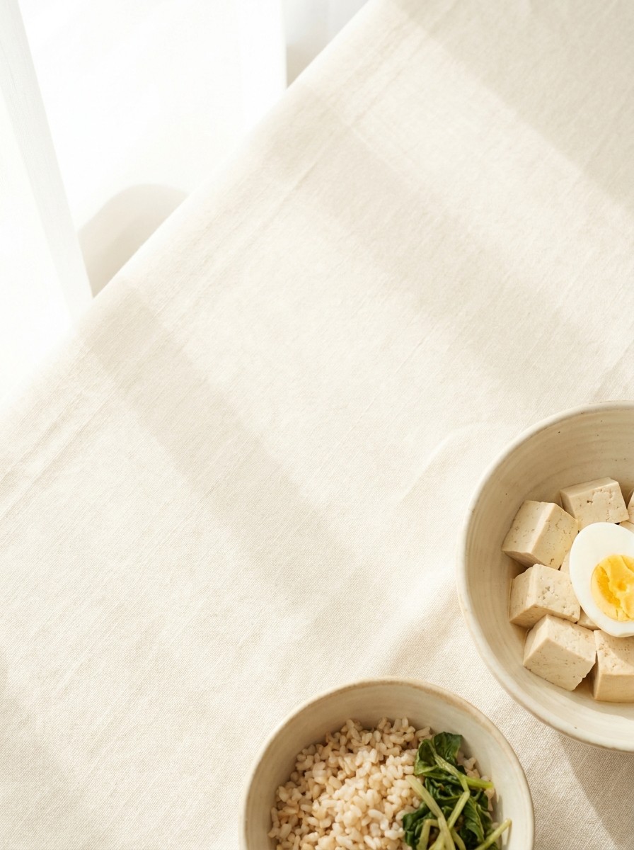 A ceramic bowl of tofu cubes with a halved boiled egg and a small bowl of brown rice with greens pushed to the lower-right of cream linen, the upper-left left empty.