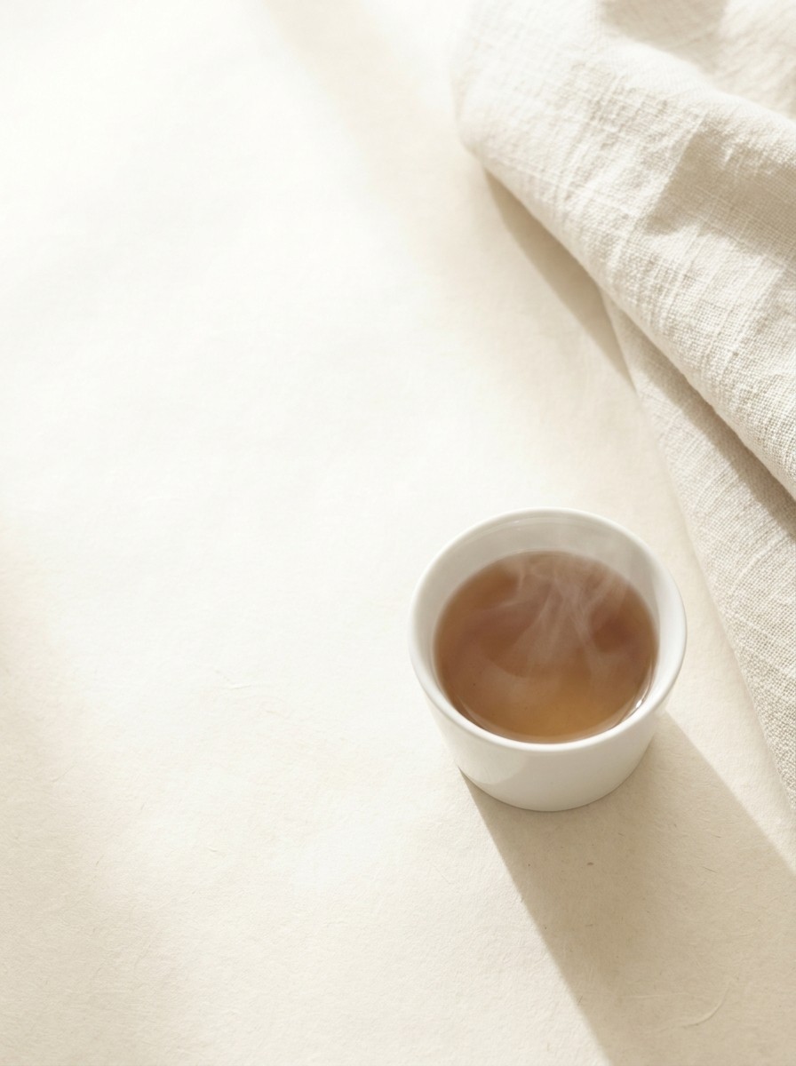 A porcelain cup of warm tea with faint steam and the corner of a linen blanket at the lower-right of cream hanji paper, the upper-left left empty.