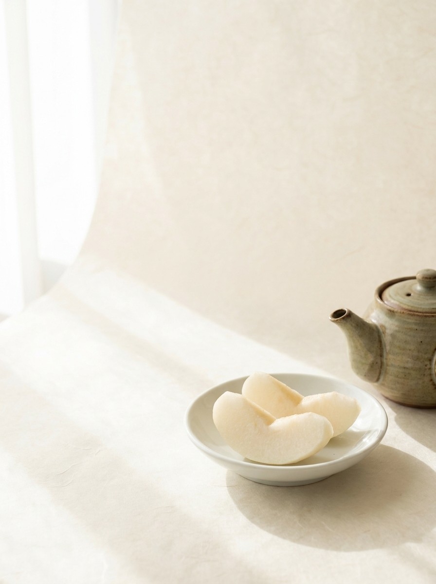 Two slices of Korean pear on a porcelain dish and a small ceramic tea pot pushed to the lower-right of cream hanji paper, the upper-left left empty.