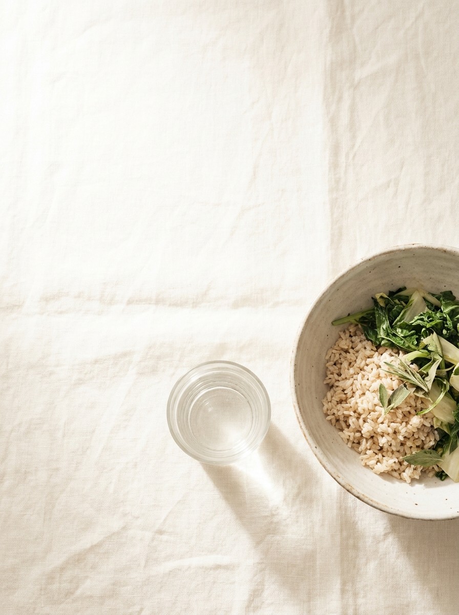 A ceramic bowl of brown rice with leafy greens and a glass of water pushed to the lower-right of cream linen, the upper-left left empty.
