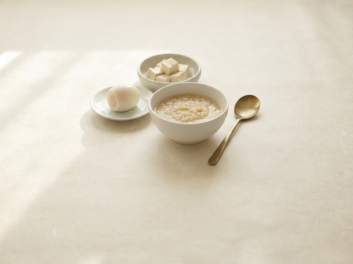 A porcelain bowl of soft rice porridge, a small dish of tofu cubes, a peeled soft-boiled egg and a brass spoon on cream hanji paper in soft window light.