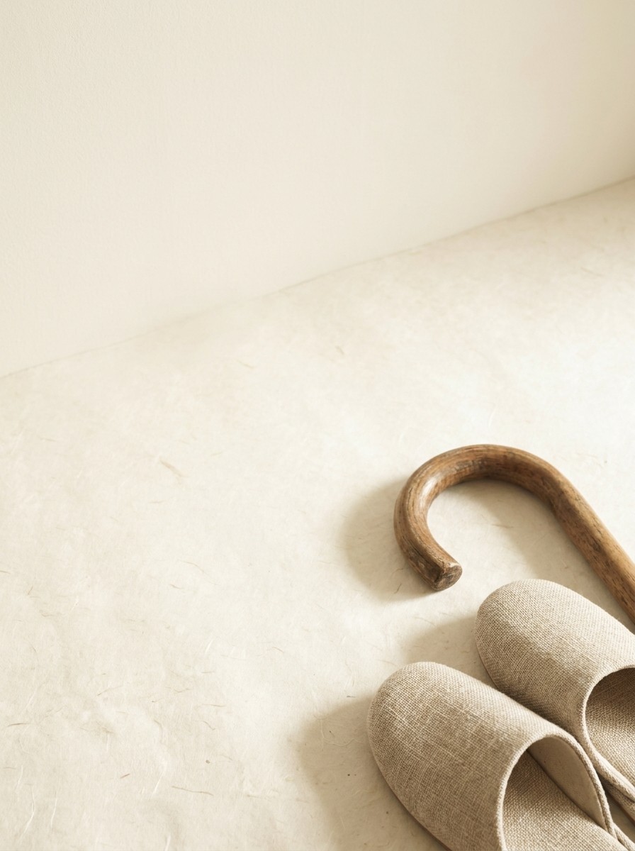 The curved wooden handle of a walking cane and a pair of soft beige slippers pushed to the lower-right of a hanji surface, the upper-left left empty.