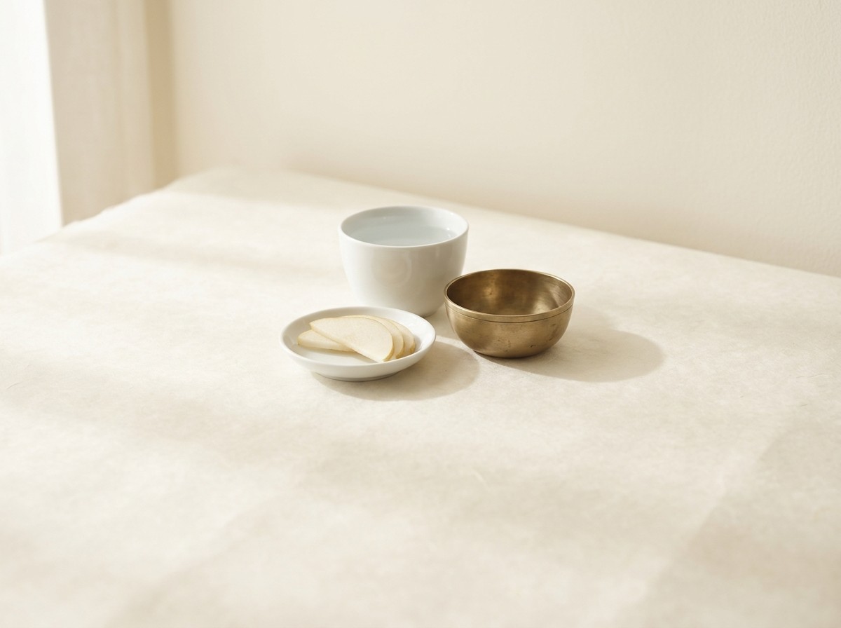 A white porcelain cup of cool water, a small dish of thinly sliced pale pear and a small brass bowl clustered on cream hanji paper in soft window light.