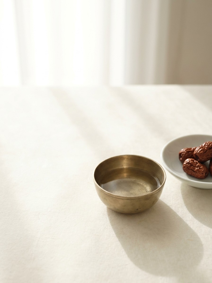 A small brass bowl of water and a white porcelain dish of dried jujubes pushed to the lower-right of cream hanji, the upper-left left empty.