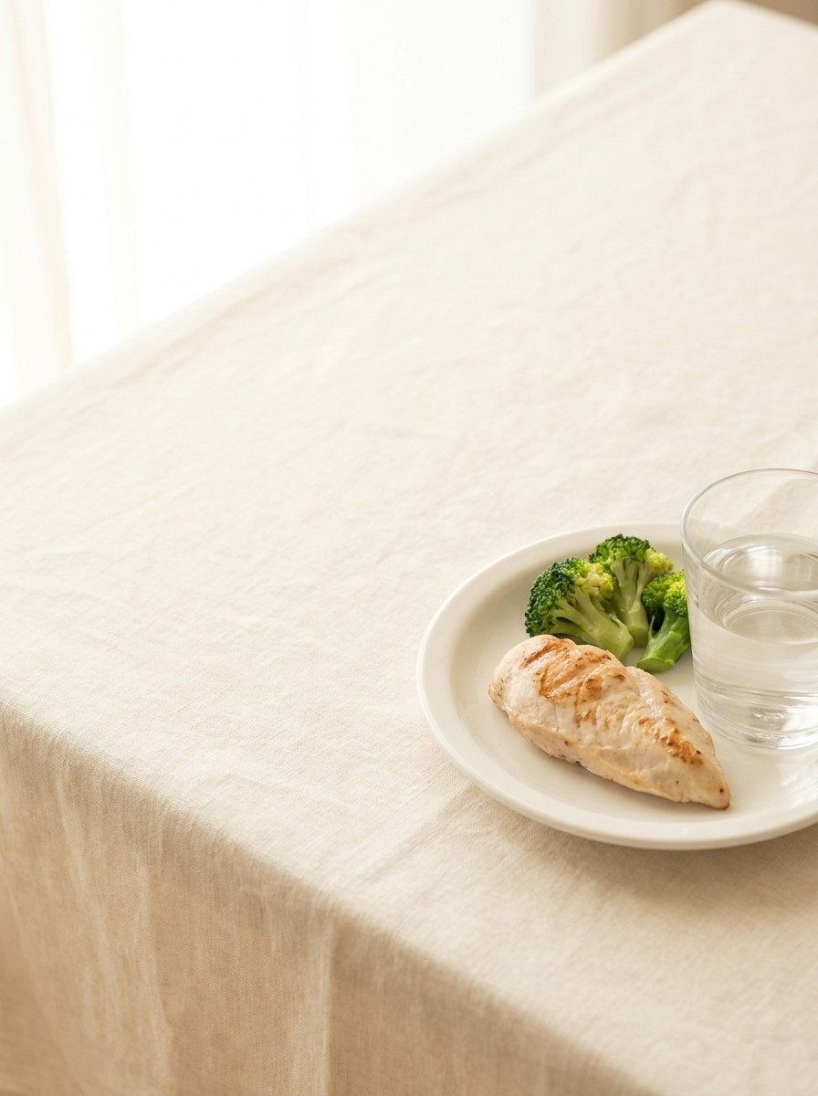 A white plate with a slice of grilled chicken breast, steamed broccoli and a glass of water at the lower-right of a cream linen tablecloth, with bright empty space at the upper-left.