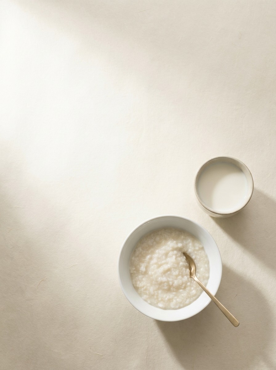 A porcelain bowl of soft rice porridge with a brass spoon and a small cup of warm milk at the lower-right of cream hanji paper, the upper-left left empty.