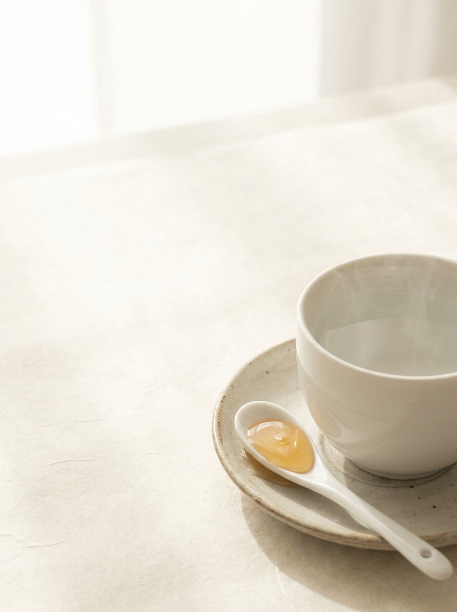 A porcelain spoon of honey on a saucer and a cup of warm water with faint steam pushed to the lower-right of cream hanji paper, the upper-left left empty.