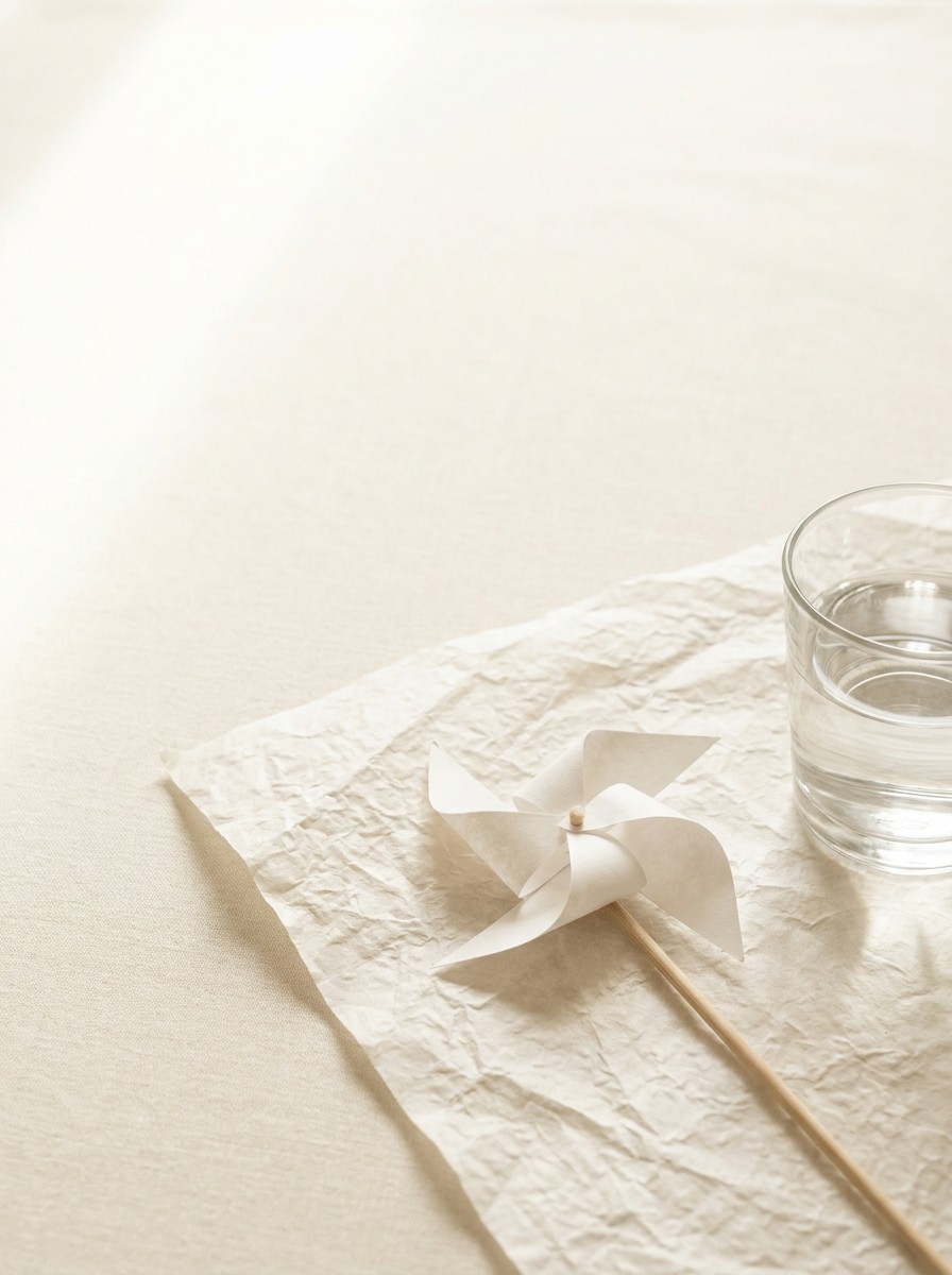 A small white paper pinwheel and a glass of water pushed to the lower-right of cream linen and hanji, the upper-left left empty in soft window light.