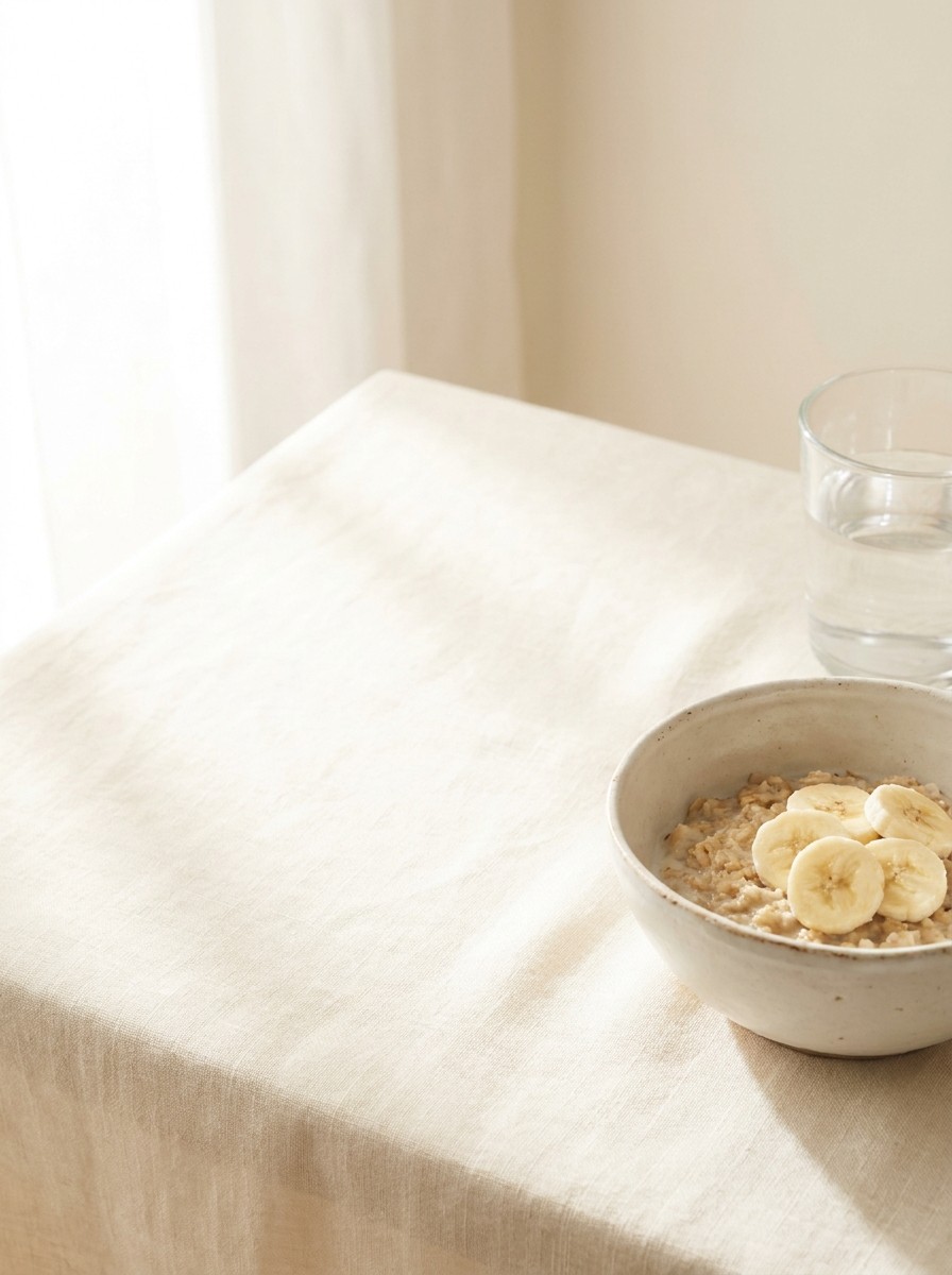 A small ceramic bowl of oatmeal with sliced banana and a tall glass of water at the lower-right of cream linen, with bright empty space at the upper-left.