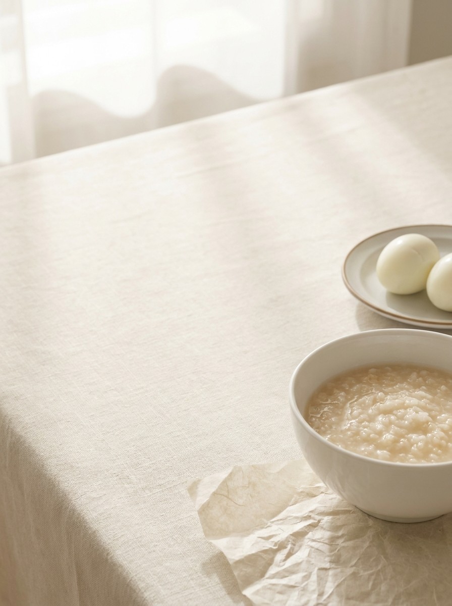 A white porcelain bowl of rice porridge and a small plate with two peeled boiled eggs pushed to the lower-right of cream linen and hanji, the upper-left left empty.