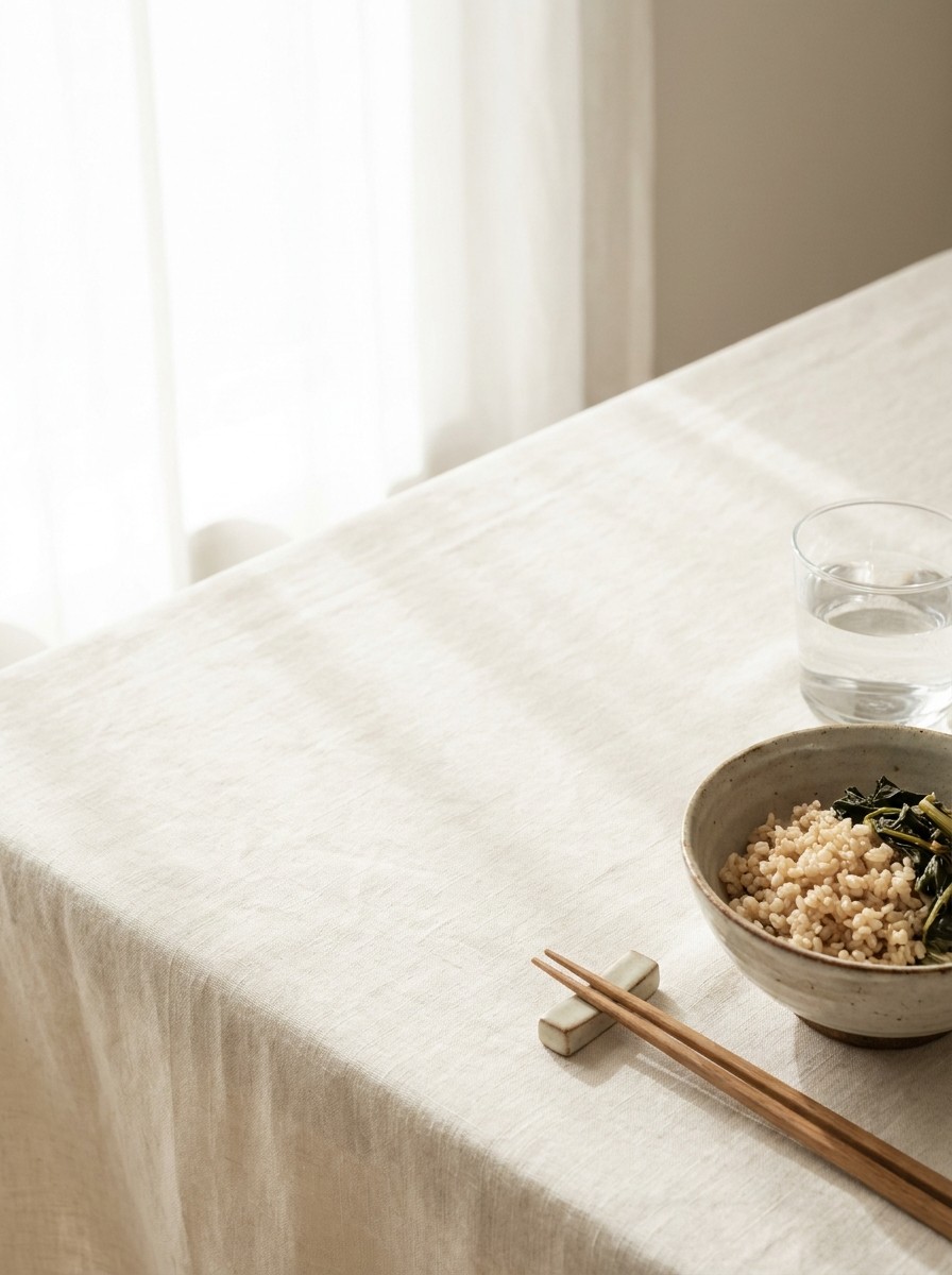 A ceramic bowl of brown rice with greens, wooden chopsticks and a glass of water pushed to the lower-right of cream linen, the upper-left left empty in soft window light.