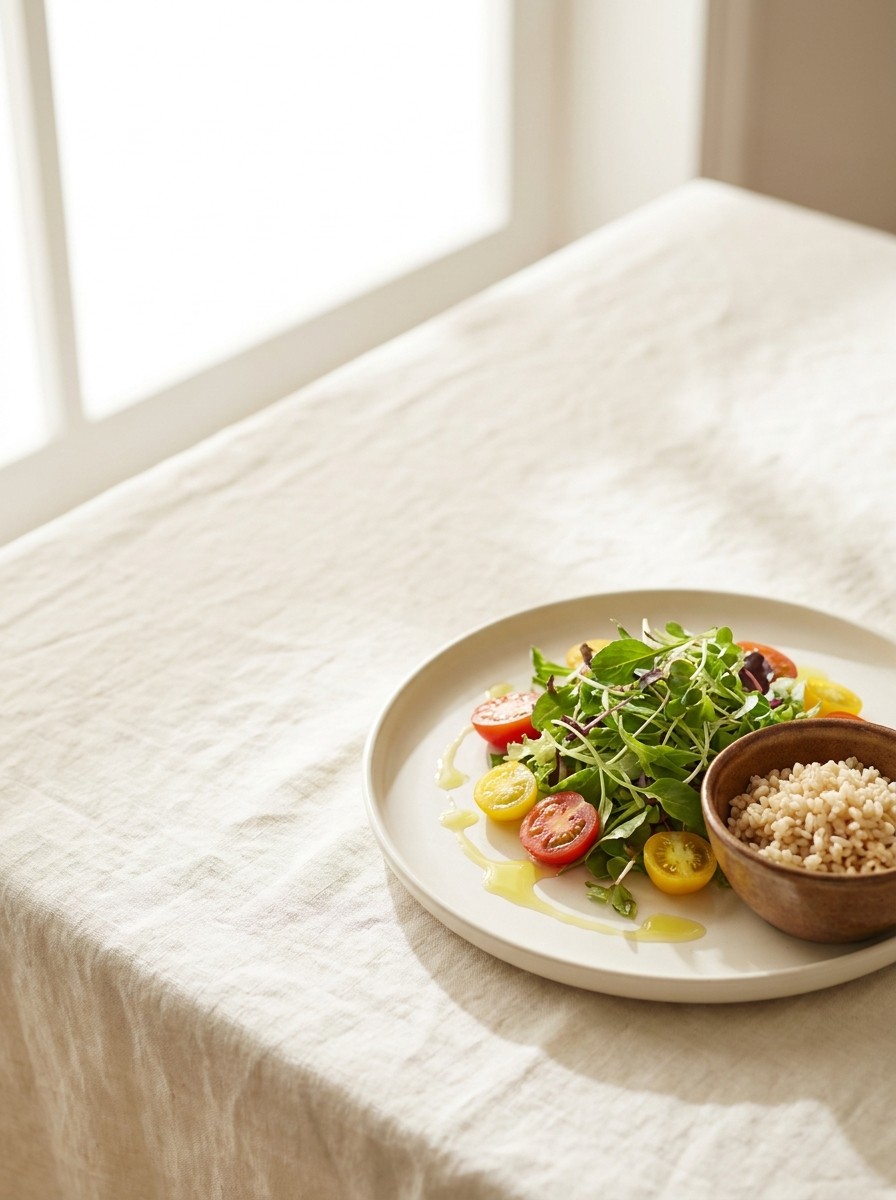 A plate of leafy greens and cherry tomatoes drizzled with olive oil and a ceramic bowl of brown rice pushed to the lower-right of cream linen, the upper-left left empty.
