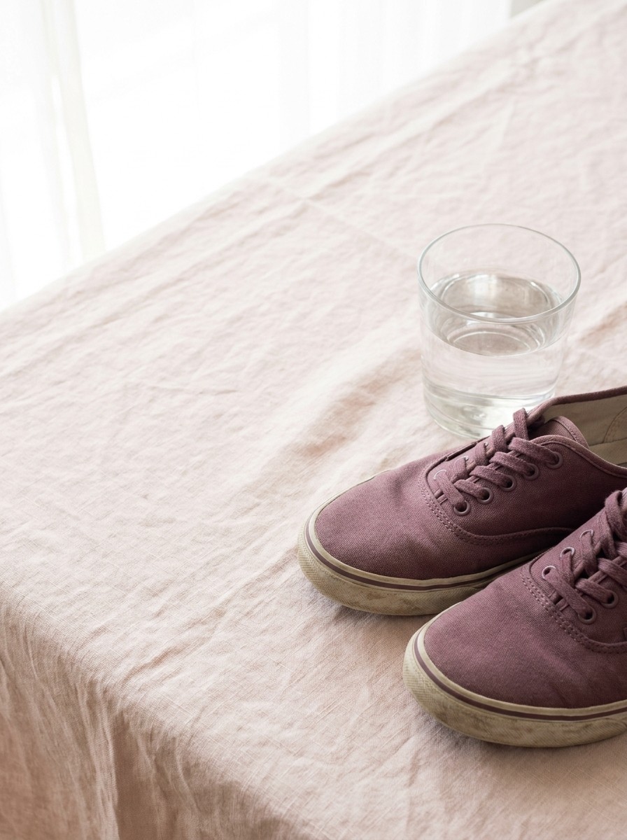 A pair of soft plum canvas walking sneakers and a clear glass of water on blush linen, with open space at upper left.