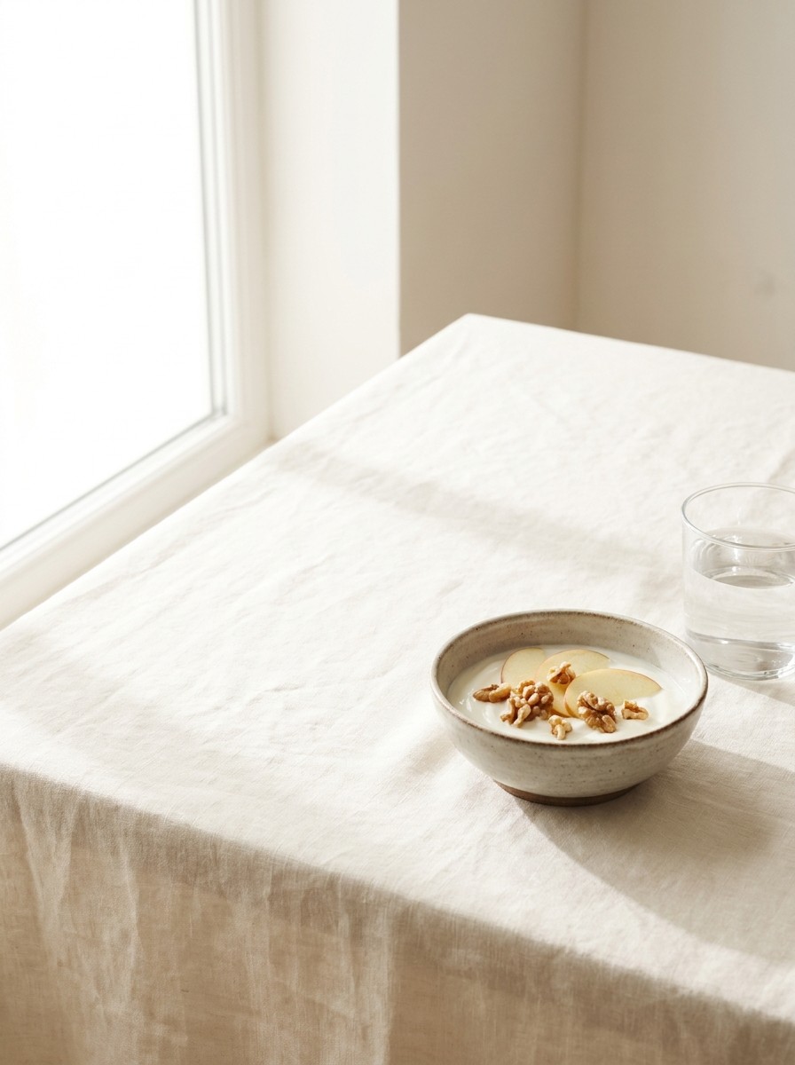A small ceramic bowl of plain yogurt with sliced apple and walnuts and a glass of water at the lower-right of cream linen, with bright empty space at the upper-left.