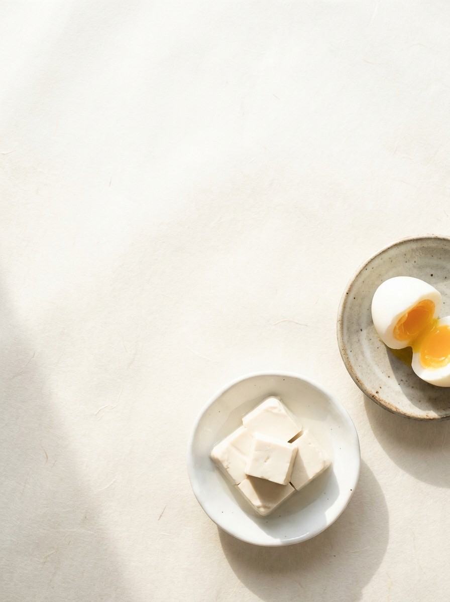 A small porcelain dish of tofu cubes and a halved soft-boiled egg on a plate at the lower-right of cream hanji paper, the upper-left left empty.