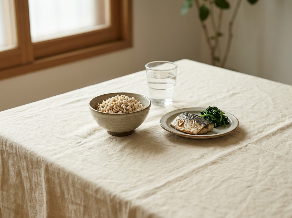 A bowl of brown rice with barley, a plate of grilled mackerel with spinach and a glass of water on cream linen in soft window light.