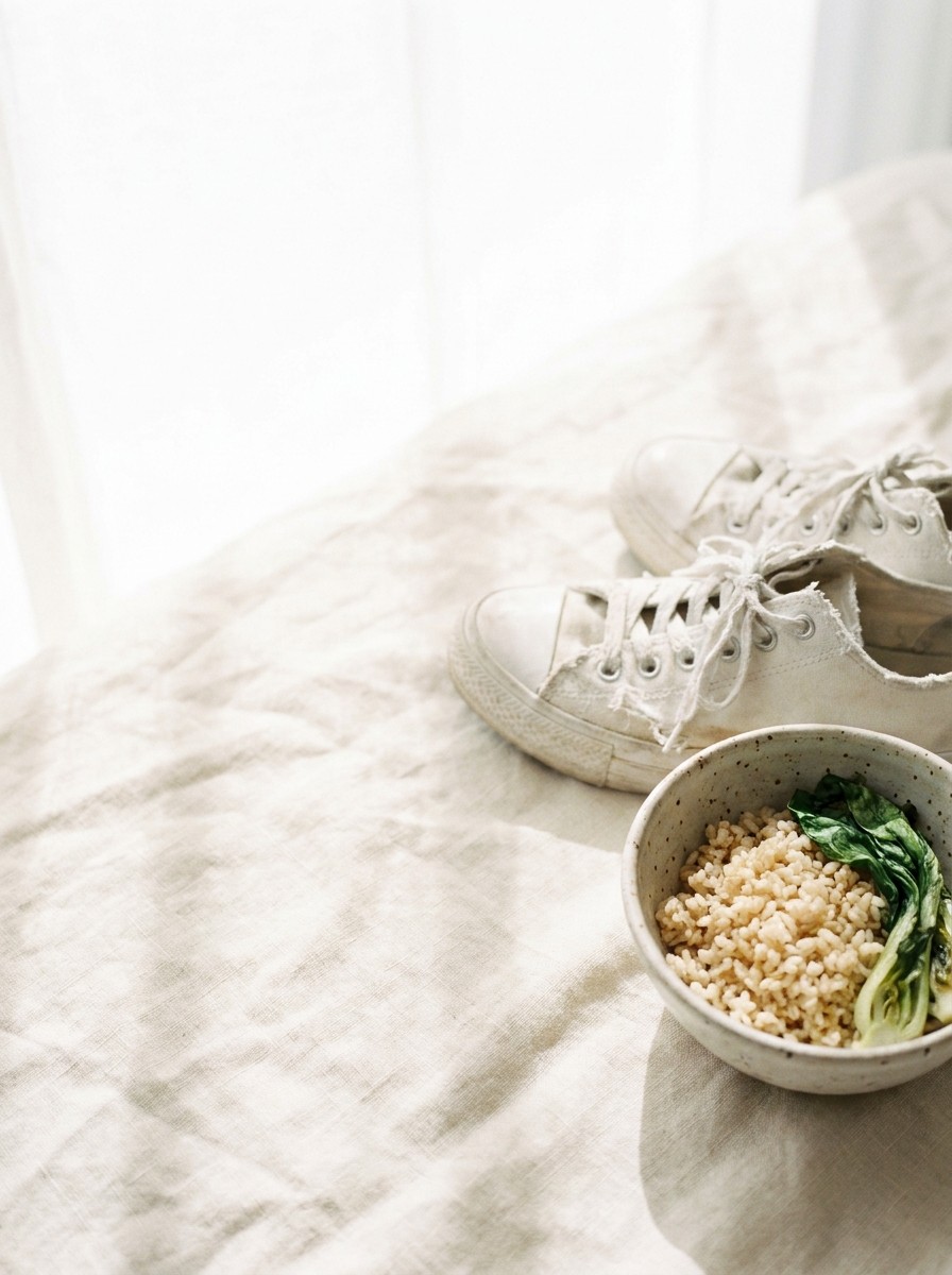 A pair of white canvas sneakers and a small bowl of brown rice with greens pushed to the lower-right of cream linen, the upper-left left empty.