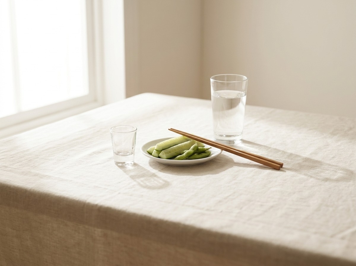 A small clear shot glass, a white plate of cucumber sticks and edamame with wooden chopsticks, and a tall glass of water clustered on a cream linen tablecloth in soft window light.