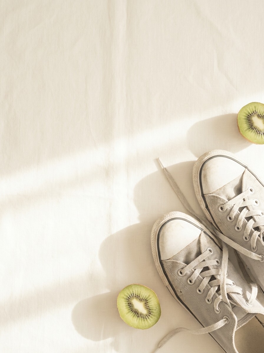 A pair of light gray sneakers and two kiwifruit halves pushed to the lower-right of cream linen, the upper-left left empty.