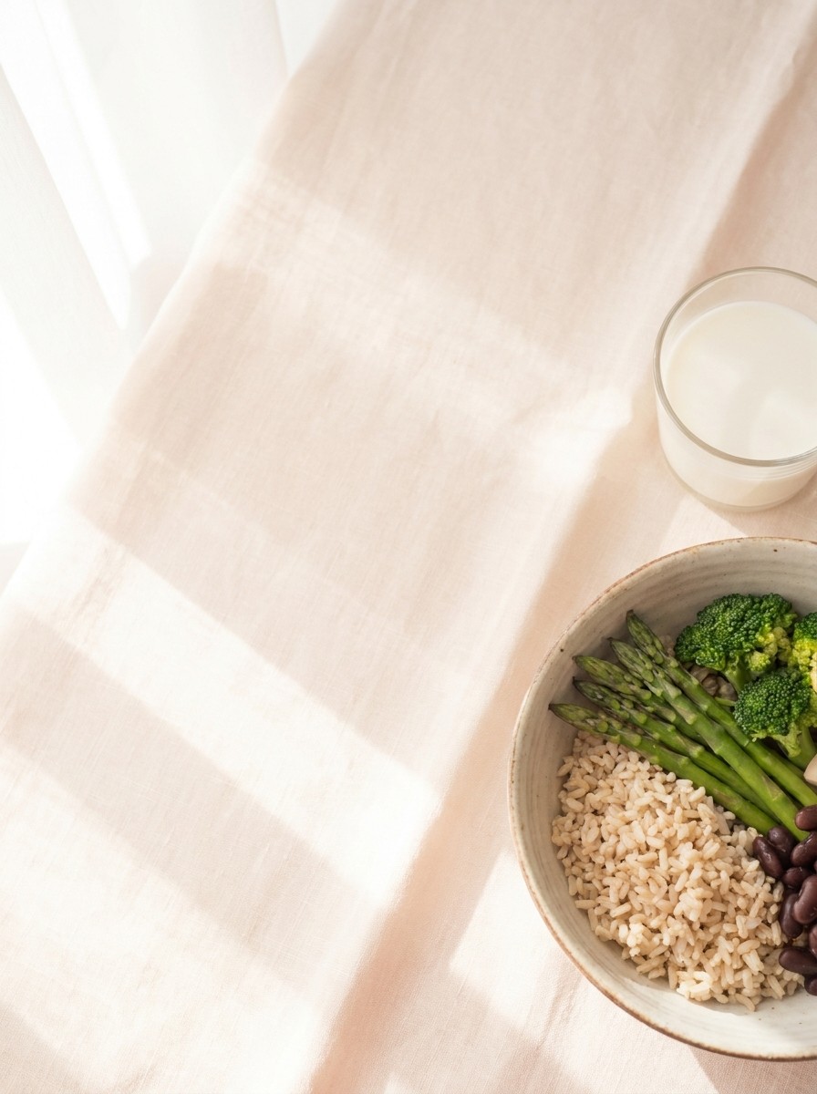 A bowl of brown rice with asparagus, broccoli and beans and a small glass of milk at the lower-right of blush-tinted linen, the upper-left left empty.
