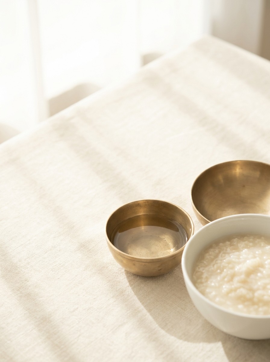 A white porcelain bowl of soft rice porridge and small brass bowls at the lower-right of cream linen, with bright empty space at the upper-left.