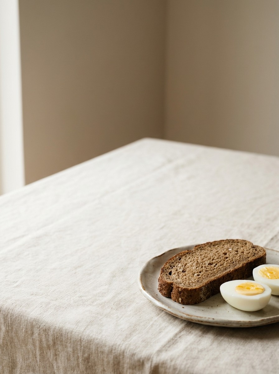 A slice of whole-grain bread and halved boiled eggs on a ceramic plate at the lower-right of cream linen, with bright empty space at the upper-left.