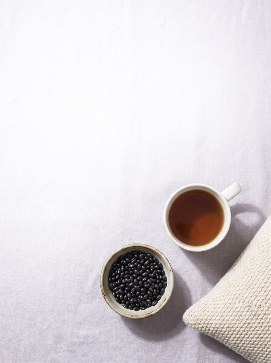 A white mug of warm tea, a small bowl of black beans and the corner of a soft knit pillow on pale lavender-grey linen, with open space at upper left.