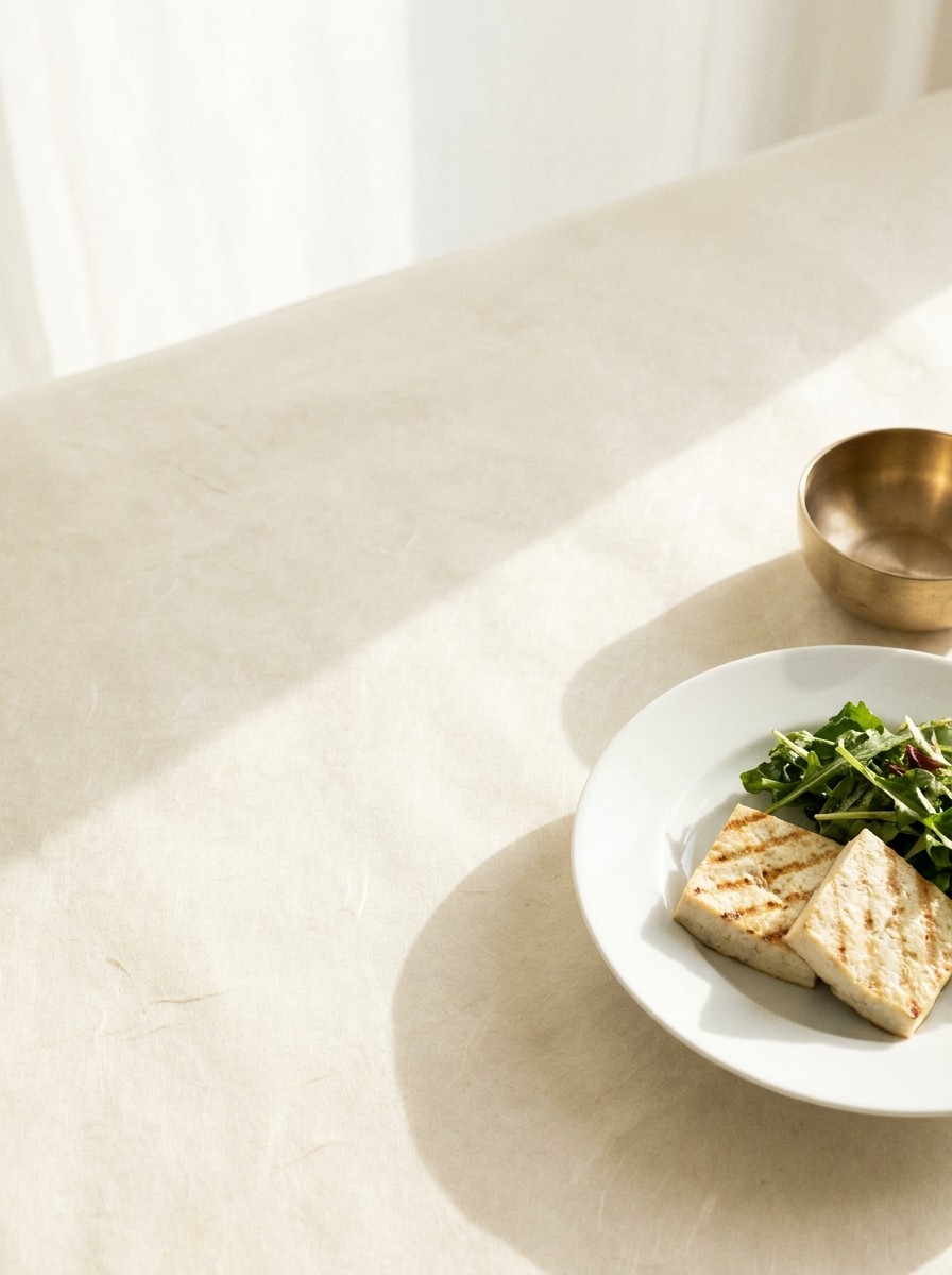 A porcelain plate of grilled tofu with leafy greens and a small brass bowl pushed to the lower-right of cream hanji paper, the upper-left left empty.