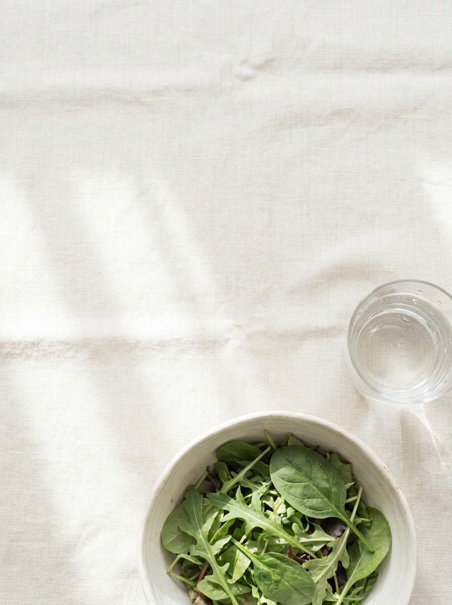 A ceramic bowl of fresh leafy greens and a glass of water pushed to the lower-right of cream linen, the upper-left left empty in soft window light.