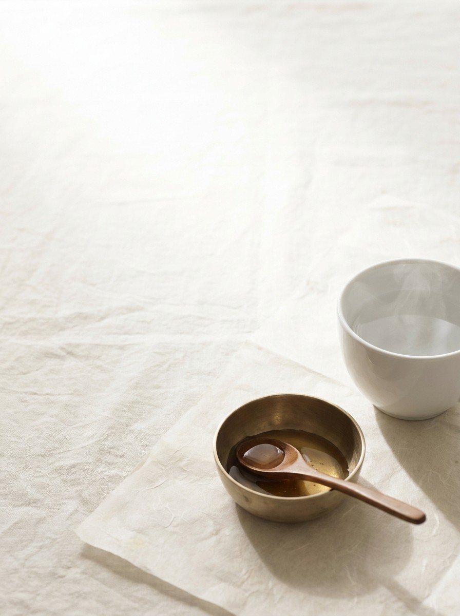 A white porcelain cup of steaming water and a brass bowl of honey with a wooden spoon pushed to the lower-right of cream linen and hanji, the upper-left left empty.