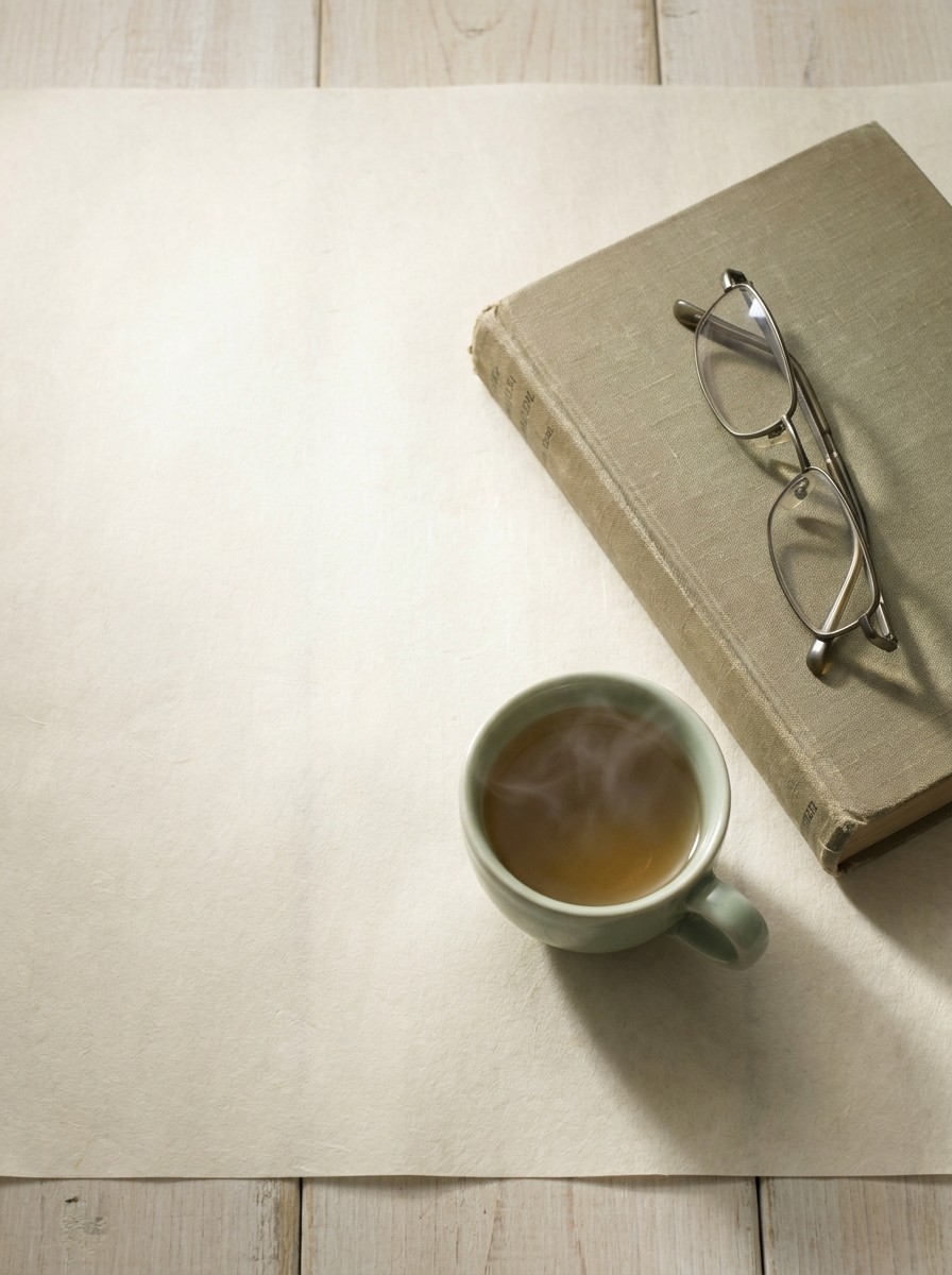 A closed cloth-bound book with folded reading glasses and a celadon cup of warm tea at the lower-right of cream hanji paper, with bright empty space at the upper-left.