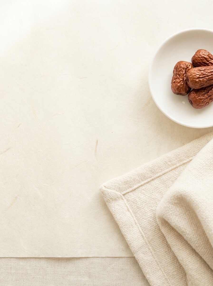 A small white porcelain dish of dried jujubes and a folded soft cotton blanket at the lower-right of cream hanji paper, with bright empty space at the upper-left.