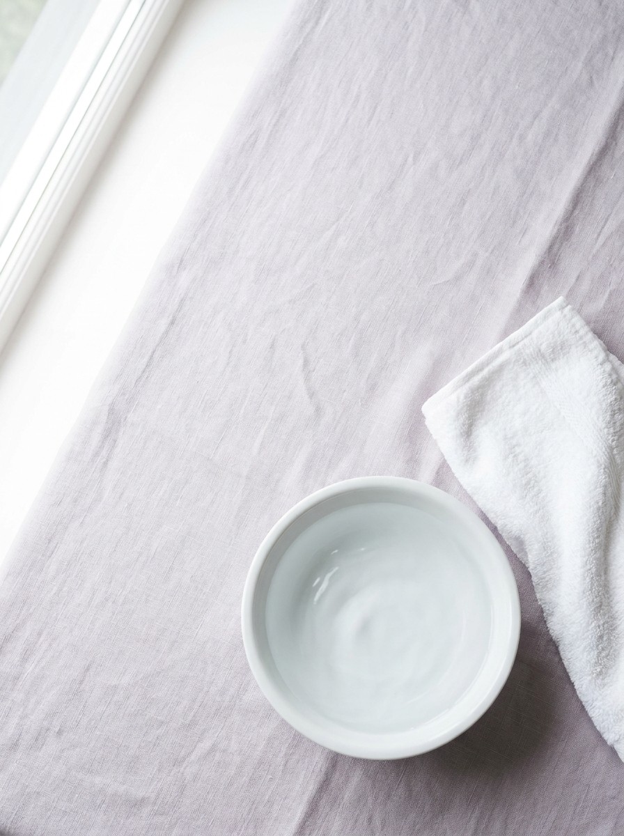 A small white ceramic basin of clear water and a folded white cotton towel on pale lavender-grey linen, with open space at upper left.