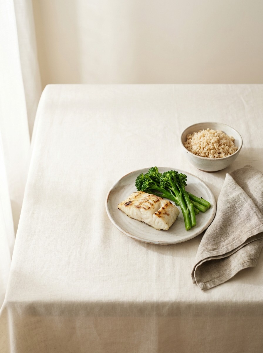 A plate of grilled white fish with steamed broccoli, a small bowl of brown rice and a folded linen napkin at the lower-right of cream linen, with bright empty space at the upper-left.