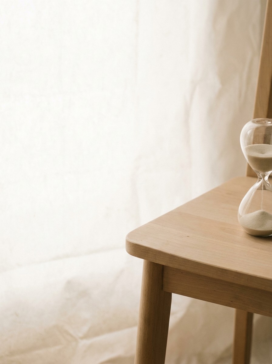 The corner of a light wooden chair seat with a small glass hourglass pushed to the lower-right of a hanji surface, the upper-left left empty.