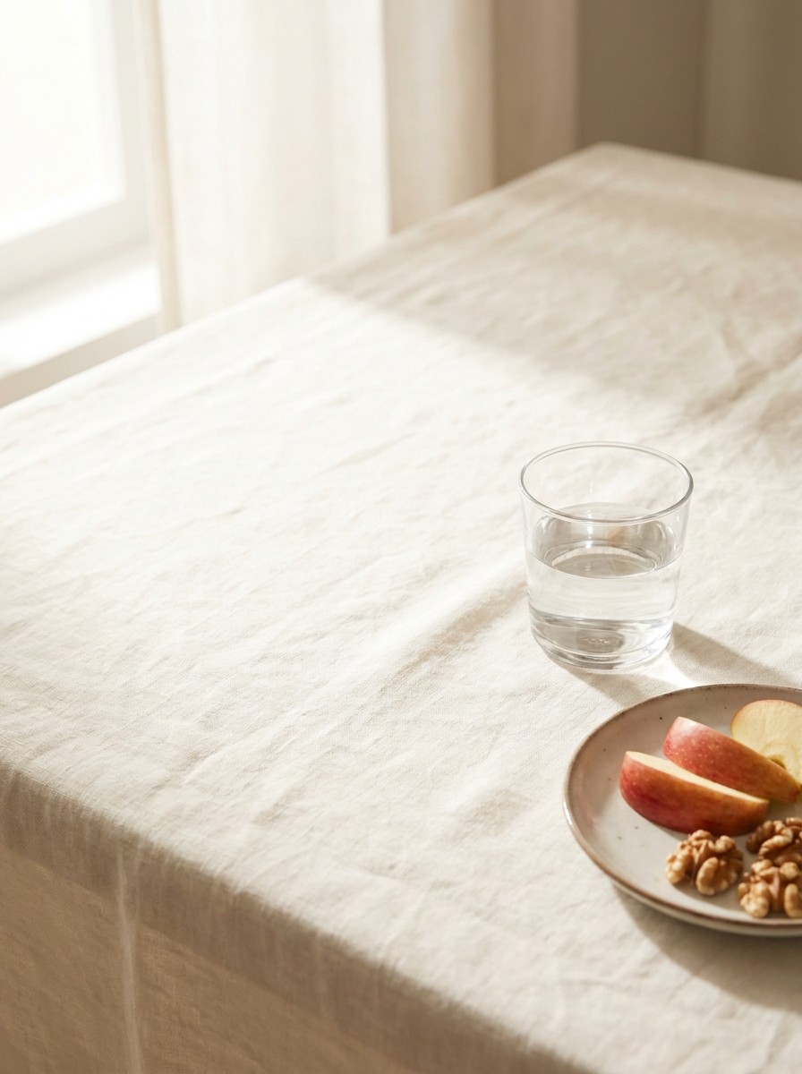 A glass of water and a small plate of sliced apple and walnuts at the lower-right of cream linen, with bright empty space at the upper-left.