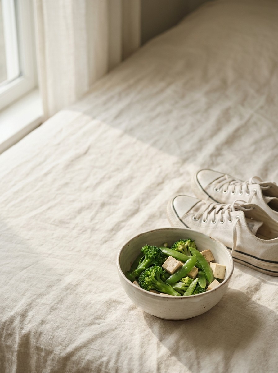 A pair of white sneakers and a ceramic bowl of steamed greens with tofu at the lower-right of cream linen, the upper-left left bright and empty.