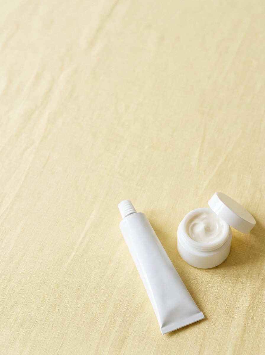 A plain white tube of ointment and a small jar of thick cream on butter-yellow linen, with open space at upper left.