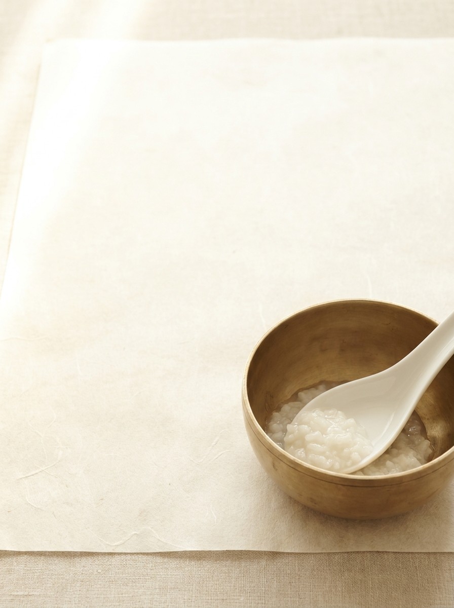 A small brass bowl of soft rice porridge with a white porcelain spoon at the lower-right of cream hanji paper, with bright empty space at the upper-left.