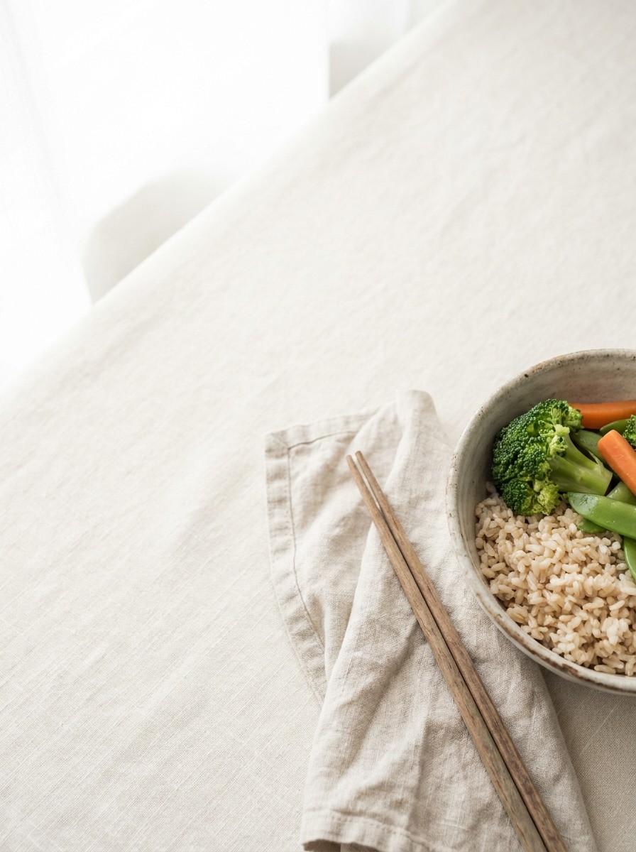 A ceramic bowl of brown rice with steamed vegetables and wooden chopsticks on a folded linen napkin at the lower-right of cream linen, with bright empty space at the upper-left.