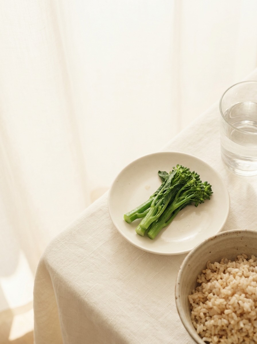 A ceramic bowl of brown rice, a small plate of steamed greens and a glass of water pushed to the lower-right of cream linen, the upper-left left empty.