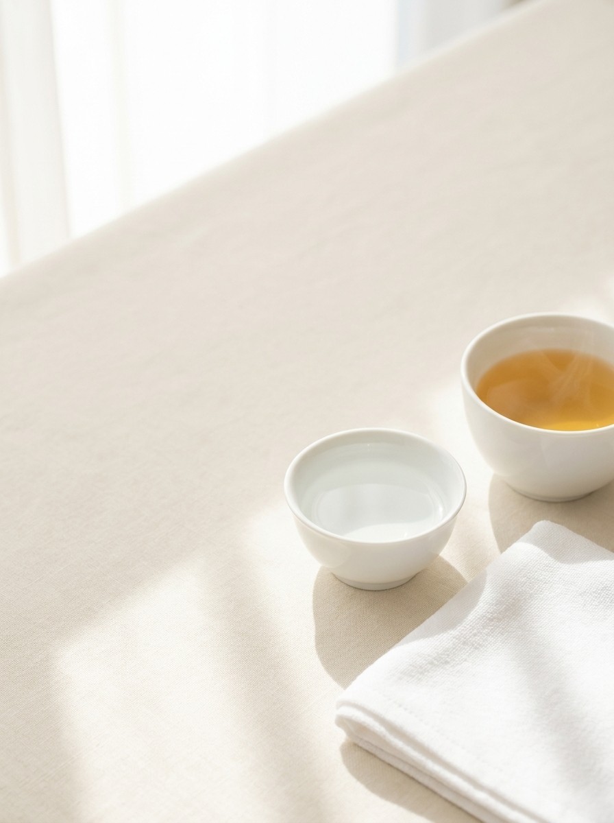 A folded white cotton towel, a small white bowl of water and a cup of warm barley tea at the lower-right of cream linen, with bright empty space at the upper-left.