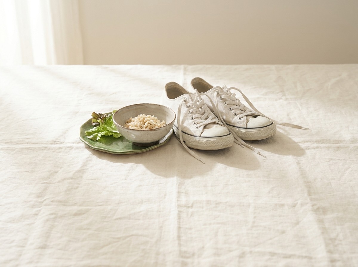 A pair of plain white canvas sneakers, a ceramic bowl of brown rice and a small green salad plate clustered on cream linen in soft window light.